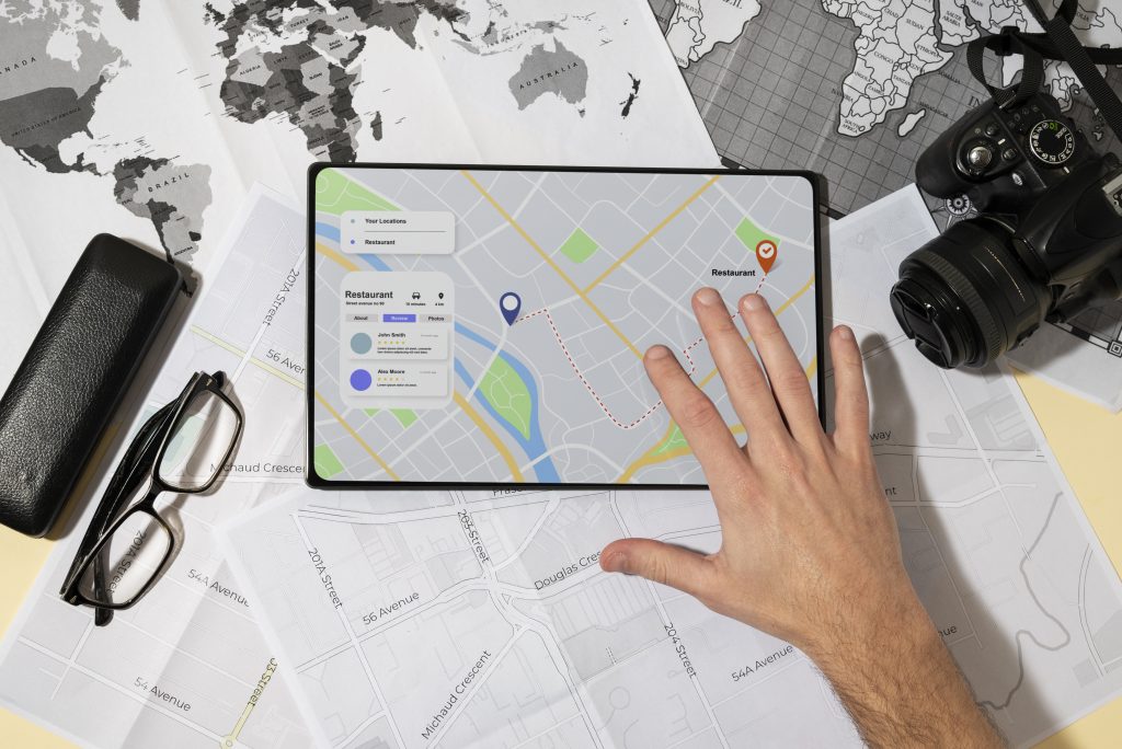 Local SEO Agency Pakistan : Hand pointing to a restaurant location on a digital map displayed on a tablet, surrounded by world and city maps, eyeglasses, and a camera.